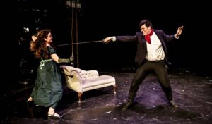 A man in a tuxedo holding a long, thin sword lunges at a woman in a green empire waist dress. She dodges the sword.