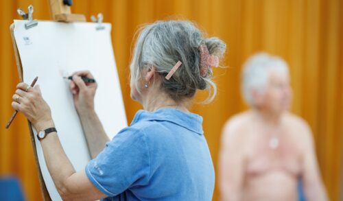 A woman in a life drawing class drawing a naked man on a large piece of paper
