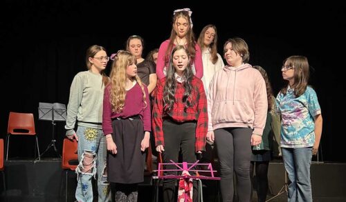 A group of young people standing in a group onstage mid performance There is a pink music stand in front of them