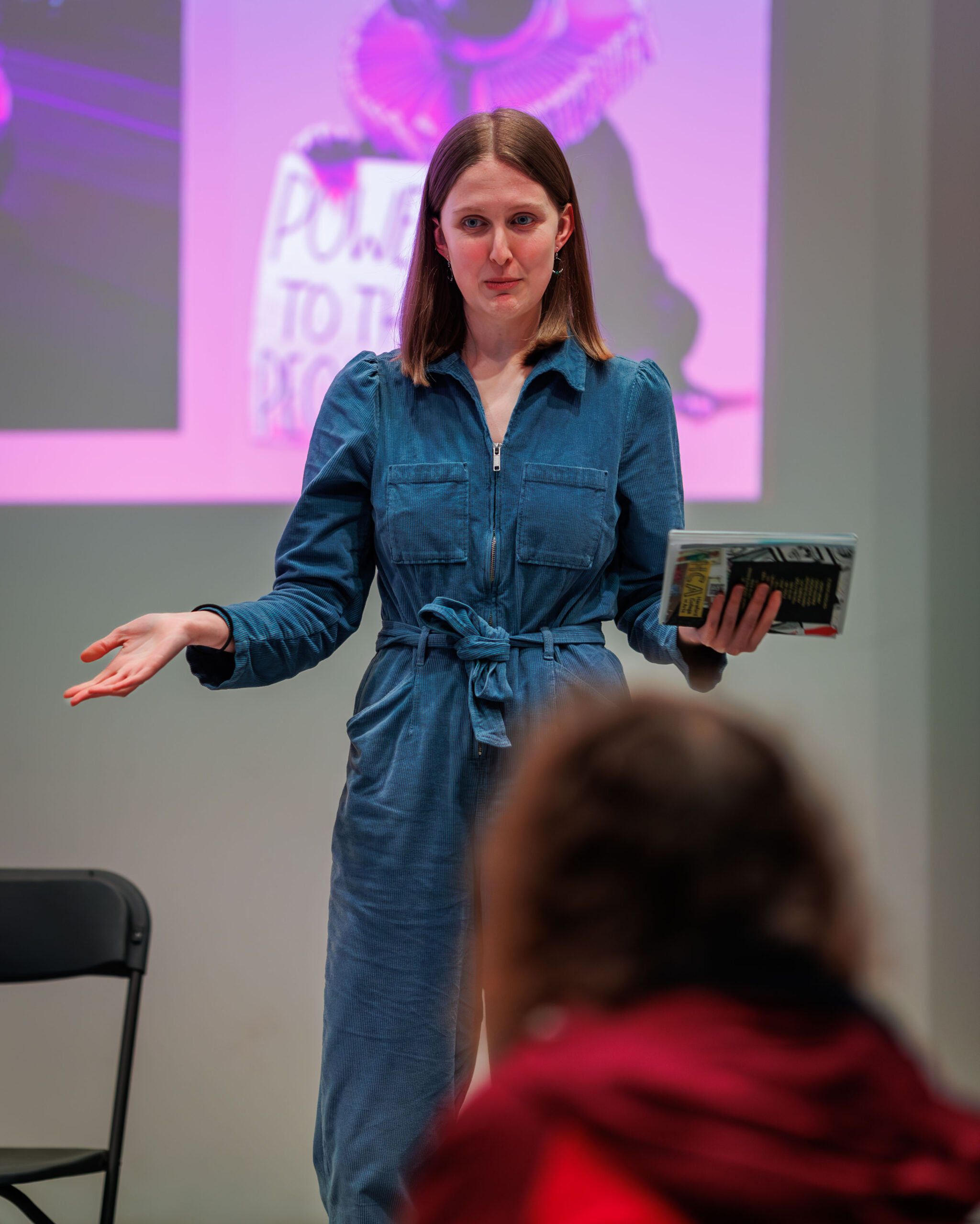 Young Film Programmer Emily stands in the Nell Gwynne Studio and presents in front of an audience. She is holding a book and is wearing a blue jumpsuit. 