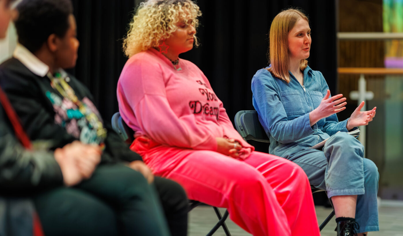 In the centre is panellist Tammy , they are dressed in a vibrant pink jumper that features the words 'In Your Dreams'. On the right is young film programmer Emily who is wearing a blue jumpsuit. Emily is speaking to the audience and is emoting with her hands. 