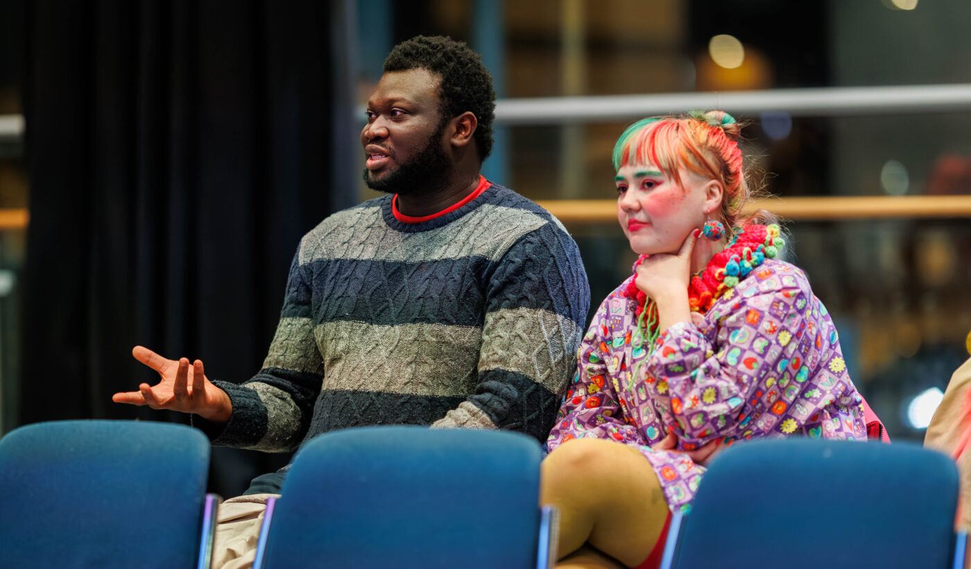 Two people sit in the audience in the Nell Gwynne. The person on the left is wearing a striped blue jumper, the person on the right is wearing a multi-coloured ruff and a vibrant top. 