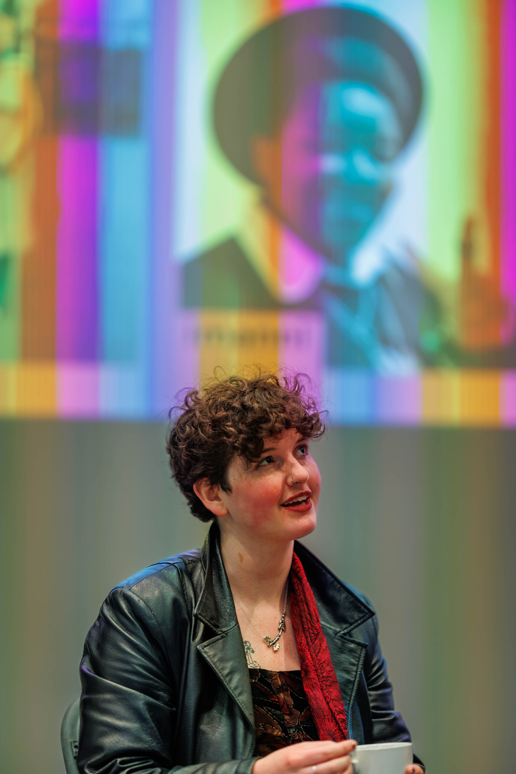 Panellist Kai sits in the Nell Gwynne Studio in a leather coat and red scarf. They are holding a mug. In the background a rainbow image is projected onto a wall. 