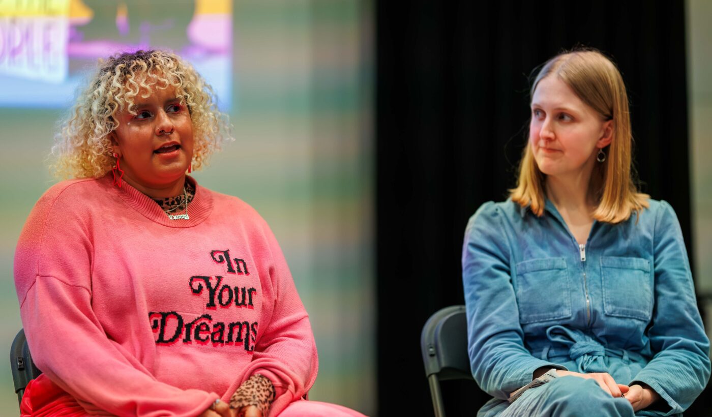 Panellist Tammy speaks to the audience, they are dressed in a vibrant pink jumper that features the words 'In Your Dreams'. On the right is young film programmer Emily who is wearing a blue jumpsuit. 
