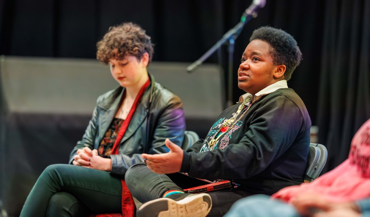 Panellist Imanni sits in the Nell Gwynne studio wearing a black jumper, jeans and sneakers. They are speaking to the audience and emoting with their hands. 