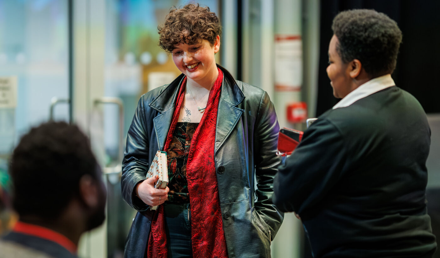 A person stands holding a notebook under their arm whilst laughing. They're wearing a leather jacket and a red scarf. 