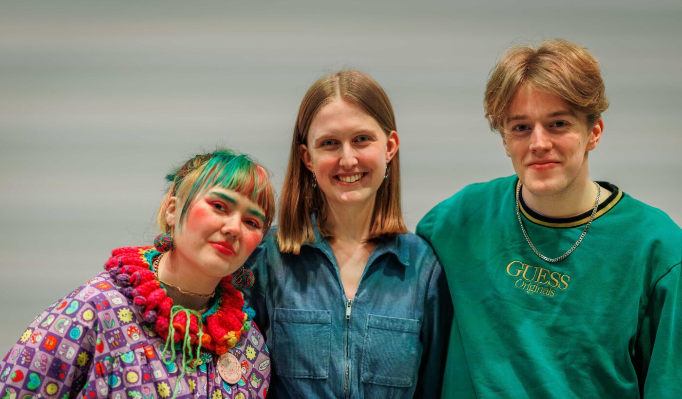 The Courtyard's Young Film Programmers stood with their arms around each other for a photo. Georgia on the left is wearing a multi-colour ruff and has green and pink hair. Emily in the middle is wearing a pink jumpsuit and has long blonde hair and Finn on the right is wearing a green jumper and has short blonde hair. 