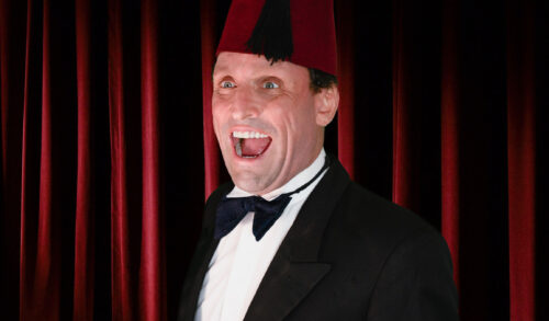An image of a man standing in a suit and bow tie wearing a red fez He smiles in front of a red curtain