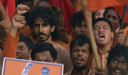 Dev Patel stands in the middle of a protest People around him are carrying orange placards