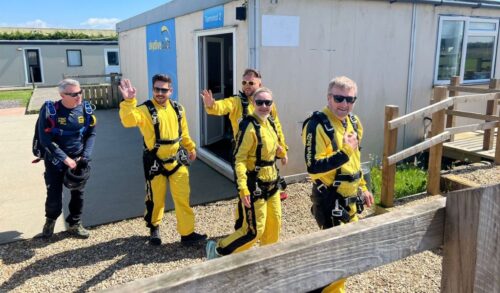 Four people in yellow boiler sky dive suits waving at the camera whilst walking