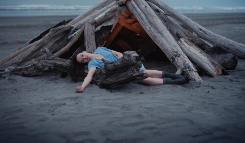 A woman lays on the ground as though she is dead Surrounding her are logs arranged to make a small structure