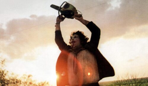 A man in a field holds a chainsaw above his head He is wearing a mask over his face