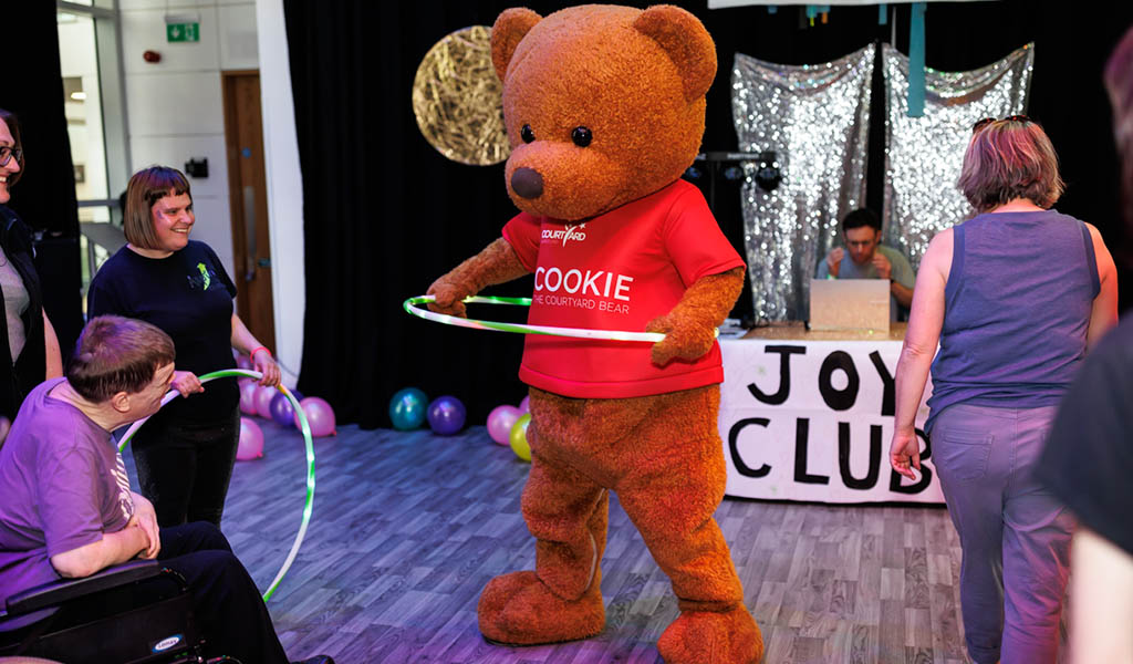 Joy Club – Inclusive Adult Rave - Cook the Courtyard Bear (a big mascot costume) does the hula hoop, whilst people watch on smiling. 