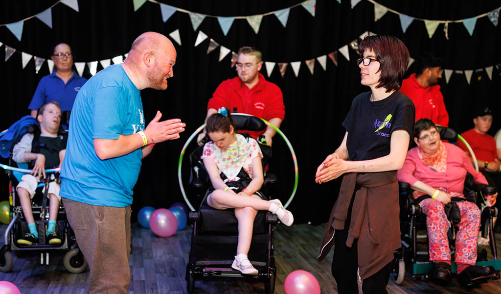 Joy Club – Inclusive Adult Rave. Three people in wheelchairs have colourful hoops around them, whilst 2 people dance in front of them. There are lots of bunting and balloons int he background. 