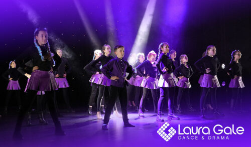 A young male performer wearing black clothing and a purple waitcoat stands centre stage surrounded by other performers who are wearing black tops with purple skirts All performers are stood with their hands on their hips