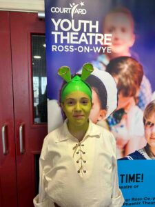 A girl dressed in a white shirt and Shrek ears stands in front of a Courtyard Youth Theatre banner