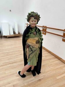 A young boy dressed up in a green robe, wearing a leaf crown and leaf face paint.