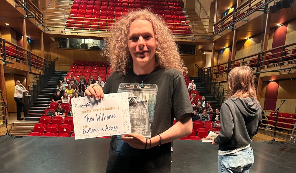 A boy stands on The Courtyard main stage holding a certificate and an award