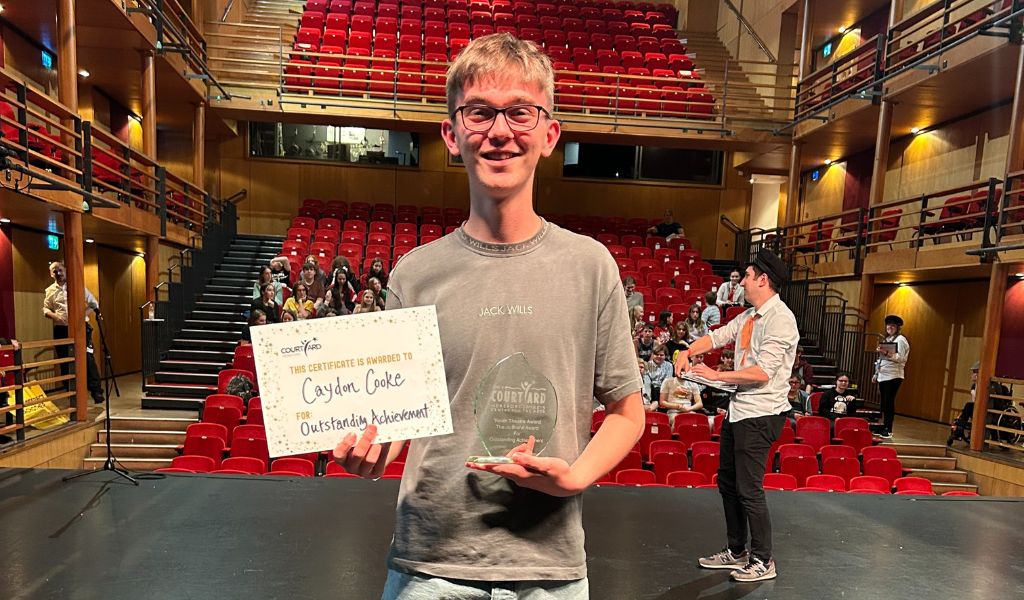 A boy stands on The Courtyard stage holding a certificate and an award