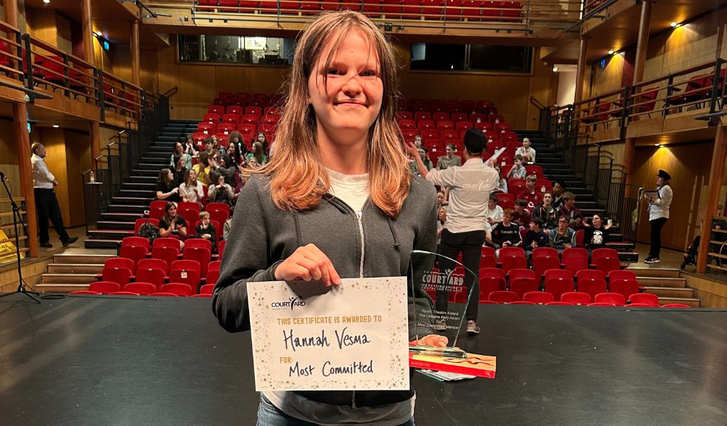 A girl stands on stage smiling, holding a certificate and an award in front of The Courtyard auditorium
