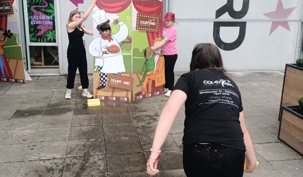 A girl in a Youth Theatre tshirt throws a wet sponge at other young people who stand behind a cardboard cut out