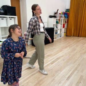 A young women stands smiling in a drama workshop next to a young girl