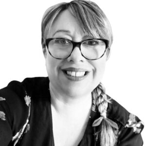 A black and white headshot of a white woman wearing glasses, with her hair tied in a plait. She is smiling at the camera