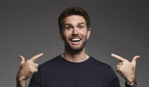 Joel Dommett stands smiling at the camera, pointing at his face