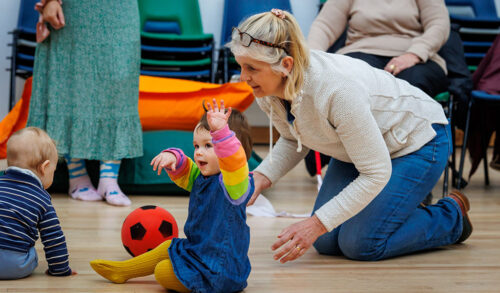A toddler wearing a rainbow jumper and denim dress sits on the floor with her hands in the air Kneeling behind her is an older woman with short blondegrey hair