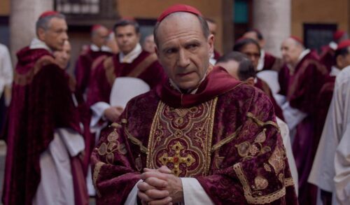 Actor Ralph Fiennes stands amongst members of the clergy in a red robe covered with gold patterns