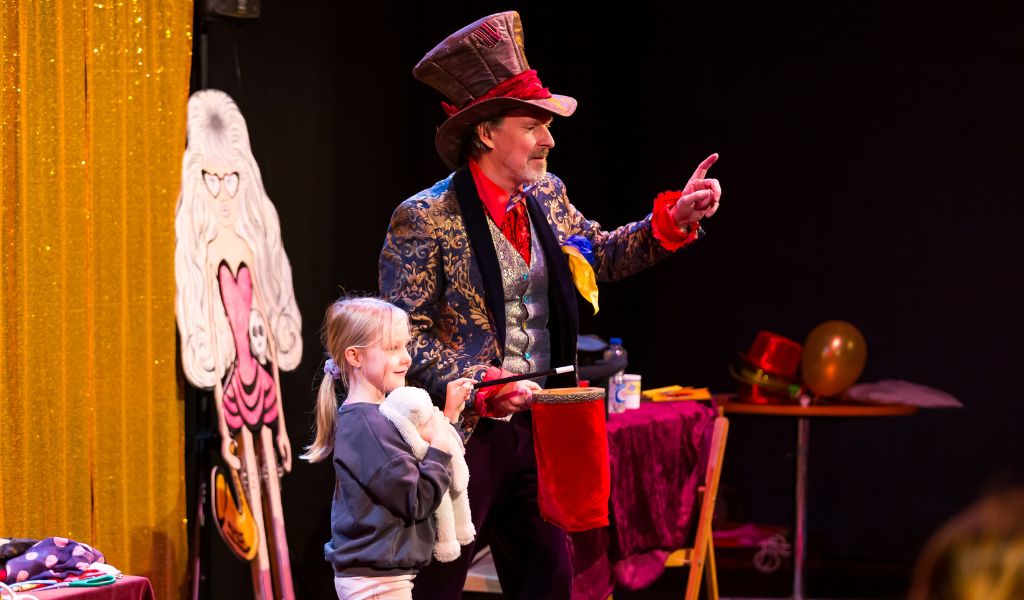 A magician performs on stage next to a little girl.