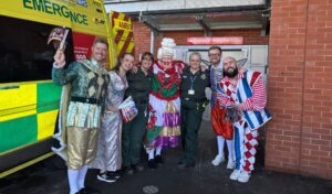 Five members of the pantomime cast in costume posing in front of an ambulance with paramedic staff