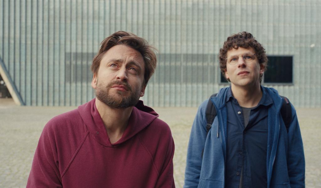 A man in a burgundy hoodie looks upward. Behind and to the right of him is a man in a blue hoodie also looking upward. 