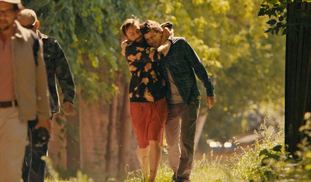 Two men walk along a path in a wooded area. One man has both his arms wrapped around the head of the other. Both men are smiling. 