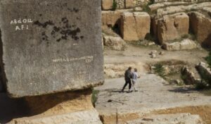 Two men walk across a stone platform which is next to a large cuboid rock. 