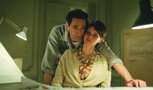 A woman sits at a desk as a man loving leans over and places his arm over her. 