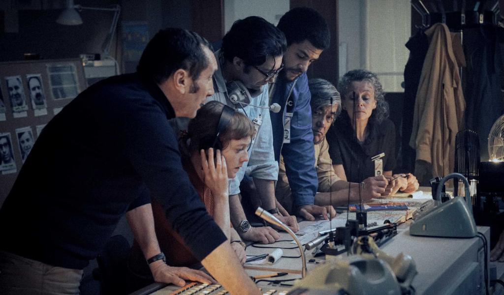A group of four men and two women gather around a control desk that is covered in dials, microphones and walkie talkies. 