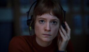 A close up image of a woman in a red sweater who is holding a pair of headphones to her head. 