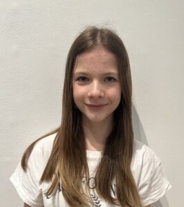 A headshot of a young girl with long brown hair, wearing a white short sleeve t-shirt.