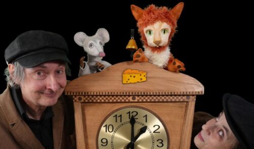 A puppet of a cat and a mouse stand on top of a grandfather clock Stood either side of the clock are people wearing black hats
