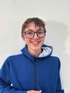 A headshot of a young boy with short brown hair. He is wearing glasses and a blue zip up hoodie and is smiling at the camera.