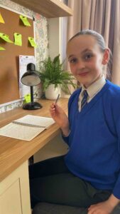 A young girl wearing a blue school uniform sits at her desk holding a pen. She is looking at the camera and smiling. 