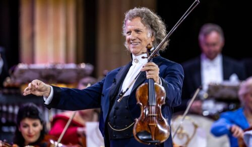 Andre Rieu stands in front of an orchestra with his hands outstretched He is holding a violin in one of his hands
