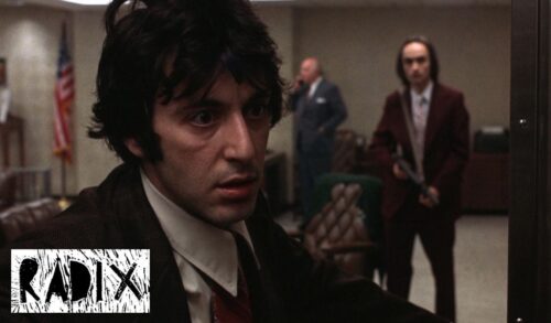 A close up image of a man in a suit and tie Behind him is another man in a suit holding a gun in front of himself