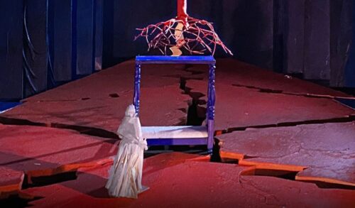 A person in a white cloak stands before the roots of a tree which are sitting on top of a blue table The floor of the stage features a number of wide cracks