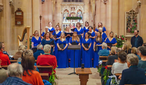 Le Voci  a female choir stand in a church all dressed in long blue dresses to an audience sat on pews