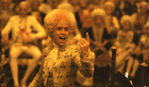 A man in an 18th century ruffled shirt and a large blonde wig stands in front of a crowd with his index finger pointing upward