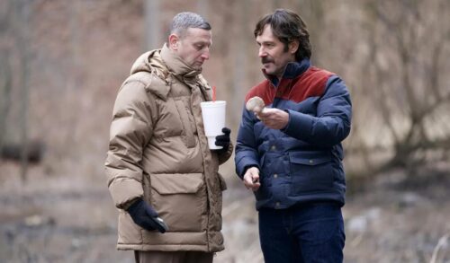 Two men in winter coats stand in a forest One man is holding a drink in a plastic cup whilst the other is holding a mushroom