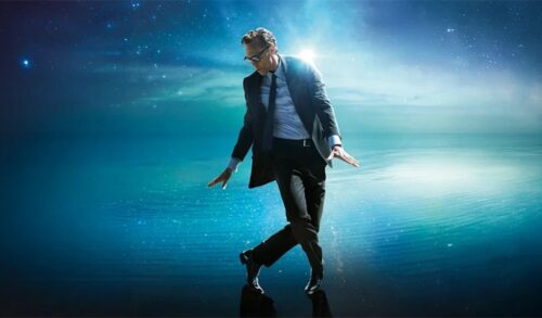 A tall man in a black suit and sunglasses dances on top of water with galaxies visible behind him