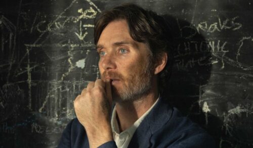 A middleaged man with midlength hair and a greying beard stands in front of a classroom chalk board He is holding his hand to his chin